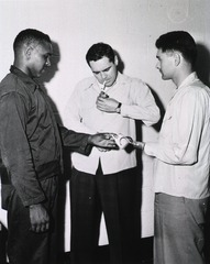 [A male amputee offers a cigarette to a second man, while a third man lights a cigarette]