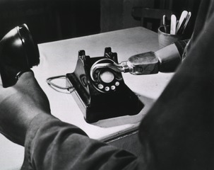 [A prosthetic hook dialing a telephone that sits on a table]