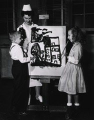 [Two children with prosthetic arms paint a picture on an easel under the supervision of a female therapist]
