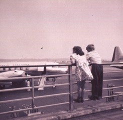 [A girl and a boy (full-length portrait, facing back) standing on the railing of an outdoor airport observation deck looking at an airplane on the tarmac] /h [graphic]