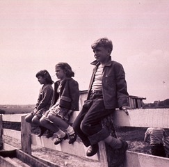 [Two girls and a boy (full-length portrait, facing left) sitting on a wooden corral (?) fence, another boy is visible in the background]