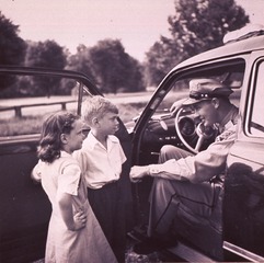 [A girl and a boy (three-quarter length portrait, facing left) standing inside the opened door of a police car talking to a Montgomery County Police officer (full-length portrait, facing left) seated in the police car]